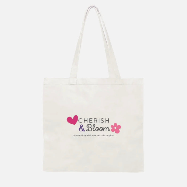 An off-white canvas tote bag with long handles, featuring a printed graphic on the front that reads 'CHERISH & Bloom' with a pink heart and flower, on a light gray background.